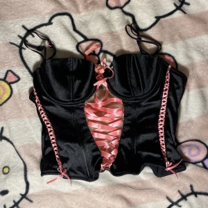 Svart korsettopp med rosa snörning H&M - Supersnygg svart korsettopp från H&M med glansig finish och rosa satinband som snörning framtill och på sidorna. Justerbara axelband och formade kupor ger en riktigt cool och feminin look. Perfekt för dig som vill sticka ut med detaljer och kontraster.