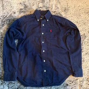 Mörkblå skjorta från Polo Ralph Lauren - S passar xs Klassisk mörkblå skjorta från Polo Ralph Lauren i bomull med vit knappstängning och röd broderad logga på bröstet. Skjortan har lång ärm, button down-krage och en tidlös passform som funkar till många olika stilar.