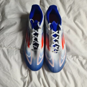 Adidas vita och blå fotbollsskor f50 - Snygga Adidas fotbollsskor i vitt med blå och röda detaljer. Skorna har snörning, spetsig tå och mönstrad ovandel i syntetmaterial. Insidan är blå och yttersulan har dobbar för bra grepp på planen. Perfekta för dig som vill sticka ut på matchen.