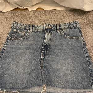 Jeanskjol från Gina Tricot - Säljer en klassisk blå jeanskjol från Gina Tricot x Izandelle. Kjolen är kort med råa kanter nertill, fem fickor och bälteshällor. Perfekt för dig som gillar en avslappnad och trendig stil.