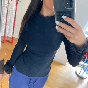 Svart långärmad topp med volang - Svart långärmad topp med volangdetaljer vid halsringning och ärmslut. Sitter tight. 