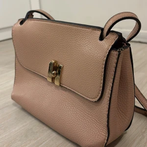 Beige axelväska från Mango i skinnimitation - Snygg beige axelväska från Mango i skinnimitation med guldigt vridlås framtill. Väskan har en stilren, boxig form och justerbar axelrem. Perfekt storlek för det viktigaste och passar till många olika outfits.