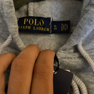 Grå hoodie från Polo Ralph Lauren - Snygg grå hoodie från Polo Ralph Lauren med dragkedja, huva och snörning. Klassisk logga broderad på bröstet och känguruficka framtill. Perfekt för en chill och stilren look. Tillverkad i mjuk bomullsmix för extra komfort.