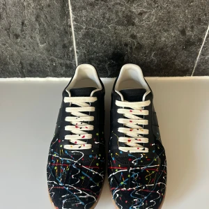 Maison Margiela sneakers med färgstänk - Unika svarta sneakers från Maison Margiela med färgstänk i rött, blått, gult, grönt och vitt. Skorna har vita snören, beige gummisula och är tillverkade i skinn. Perfekt för dig som vill sticka ut med en konstnärlig vibe. Box följs med samt tveka inte på att höra av dig!
