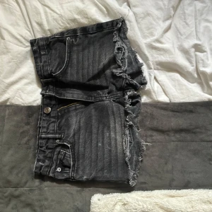 Svarta jeansshorts från Gina Tricot - Snygga mörkgråa  jeansshorts från Gina Tricot young med råa, fransiga kanter och klassisk femficksdesign. Shortsen har dragkedja och knapp framtill samt bälteshällor. Perfekta för en chill sommarlook. Lågmidjade