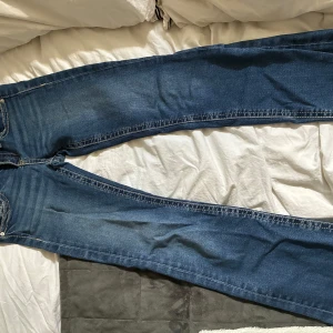 Blå bootcut jeans med fickdetaljer - Snygga blå jeans med bootcut passform och klassiska kontrastsömmar. Baksidan har två fickor med lock och knappdetaljer. Jeansen har en något utsvängd benform och är tillverkade i slitstarkt denimtyg. Spruckna på baksidan. Därför billigt pris. Priset går att diskuteras. Väldigt lågmidjade och bootcut.