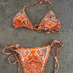 Orange mönstrad bikini med knyt - Snygg orange bikini med paisley-inspirerat mönster i vitt, rött och brunt. Klassisk trekantsmodell upptill och trosa med höga skärningar och knyt i sidorna. Perfekt för stranden eller poolen, ger riktigt somriga vibes! 🥰