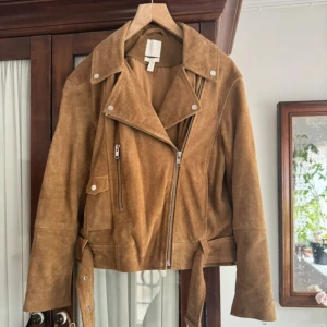 Brun mocka bikerjacka med bälte - Snygg bikerjacka i brun mocka från H&M med klassisk sned dragkedja, bred krage och bälte i midjan. Oversized 