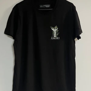 Svart Amiri t-shirt med ängeltryck - Svart t-shirt från Amiri med stort tryck av en ängel och gröna blad på ryggen samt mindre tryck på bröstet. Klassisk rund hals och korta ärmar. Skön passform och tillverkad i mjuk bomull. Perfekt för dig som gillar streetwear med unik design.