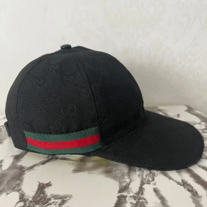 Guccu keps  - Snygg svart keps från Gucci med diskret GG-monogram över hela tyget. Klassisk böjd skärm och ikoniska gröna och röda ränder på sidan. Justerbar baktill för perfekt passform. Perfekt accessoar för en trendig look. Skriv för mer info! 