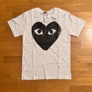 Vit Comme des Garçons Play t-shirt - Snygg vit t-shirt från Comme des Garçons Play med det ikoniska svarta hjärtat med ögon tryckt på bröstet. Klassisk rund halsringning och korta ärmar. Tillverkad i mjuk bomull, perfekt för en clean och trendig look.