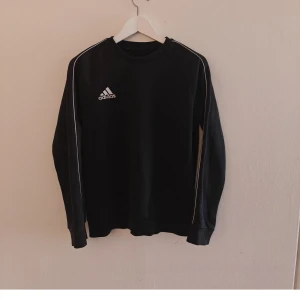 Adidas sweatshirt  - Svart långärmad sweatshirt från Adidas med broderad logga på bröstet och vita ränder längs ärmarna. Klassisk crewneck-modell i mjuk bomullsblandning, perfekt för en sportig och avslappnad stil.