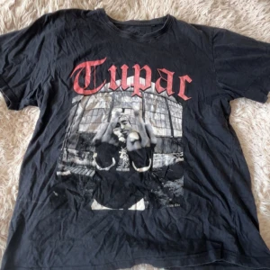 Svart Tupac t-shirt med tryck - Svart t-shirt med stort tryck av Tupac på framsidan och röd text. Klassisk passform och rund halsringning. Perfekt för dig som gillar streetwear och musikikoner.