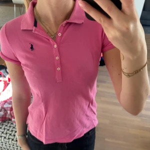 Rosa pikétröja från Ralph Lauren - Säljer en klassisk rosa pikétröja från Ralph Lauren med korta ärmar och knappar framtill. Tröjan har den ikoniska broderade loggan i marinblått på bröstet.