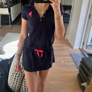 Mörkblå pikéklänning från Ralph Lauren - Mörkblå pikéklänning från Ralph Lauren med korta ärmar, klassisk krage och knappar framtill. Stor rosa logga på bröstet och siffra på ärmen. Midjan har ett rosa knytband som detalj.