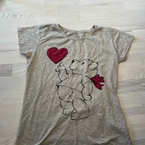 Beige t-shirt med björntryck från Love to Dream - Beige t-shirt med ett stort tryck av en björn som håller i ett rött hjärta. T-shirten har korta ärmar och en rund halsringning. Perfekt för dig som gillar gulliga motiv och vill ha en avslappnad stil.