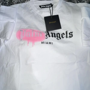 Vit Palm Angels t-shirt med Miami-tryck - Säljer en vit t-shirt från Palm Angels med svart och rosa Miami-tryck på bröstet. T-shirten har klassisk passform och är tillverkad i polyester. Perfekt statement-plagg med cool streetkänsla.   Skick 10/10✅