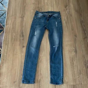 Blå skinny jeans med slitningar - Säljer ett par blå skinny jeans med slitningar på framsidan och klassiska bakfickor. Jeansen har en normal passform i midjan och är tillverkade i denim med lätt stretch. Perfekta för en avslappnad och trendig look.