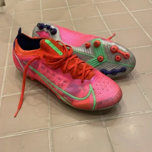 Nike Mercurial fotbollsskor i neonrosa och grönt - Säljer ett par Nike Mercurial fotbollsskor i neonrosa med gröna detaljer och röda skosnören. Skorna har låg profil, rund tå och platt sula med dobbar för bra grepp på planen. Perfekta för dig som vill sticka ut på fotbollsplanen! Skick 7,5/10 det som drar ner den en 10 är att dem e lite använda men inget som påverkar spelandet bara utseendet på nära håll! Skriv om du e intresserad 