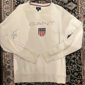 Vit sweatshirt från GANT med broderad logga - Vit sweatshirt från GANT med broderad logga och sköld på bröstet. Tröjan har rund halsringning och långa ärmar. Klassisk och stilren design med ribbade muddar vid ärmslut och nederkant.