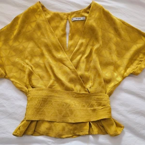 Gul omlottopp från Mango - Säljer en gul omlottopp från Mango med bred midjedel och korta ärmar. Toppen har subtilt geometriskt mönster i tyget och en snygg v-ringning framtill. Perfekt för att liva upp garderoben med lite färg!