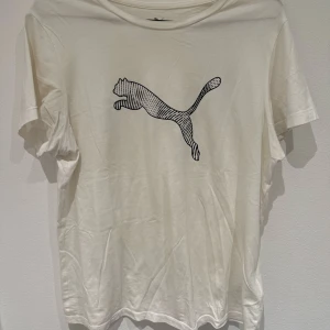 Vit t-shirt från Puma med logga - Vit t-shirt från Puma med klassisk svart logga framtill. T-shirten har rund halsringning och korta ärmar. Perfekt för en sportig och avslappnad stil.