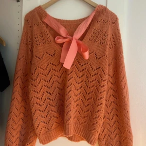 Stickad orange tröja med rosett från NLY Trend - Superfin stickad orange tröja från NLY Trend med hålmönster och en dekorativ rosett i halsen. Tröjan har lång ärm och en lätt oversized passform. Perfekt för att liva upp garderoben med lite färg!