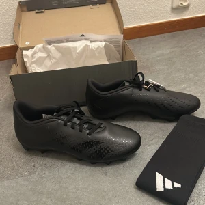 Adidas Predator Accuracy.4 fotbollsskor - Säljer ett par svarta Adidas Predator Accuracy.4 fotbollsskor med snörning. Skorna har en diskret mönstrad yta och klassisk design med Adidas-logga på sulan. Perfekta för dig som vill ha stilrena och funktionella skor på planen. Helt oanvända och i mycket bra skick