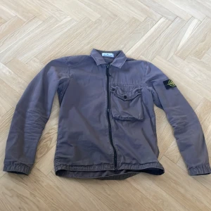 Grå vindjacka från Stone Island - Säljer en grå vindjacka från Stone Island med klassisk logotyp på ärmen. Jackan har dragkedja framtill, en bröstficka med knapp och krage. Perfekt för dig som gillar stilrena och sportiga plagg.