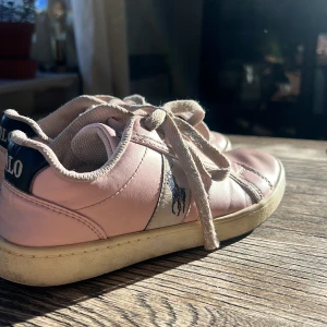 Rosa sneakers från Polo Ralph Lauren - Säljer ett par ljusrosa sneakers från Polo Ralph Lauren med vita snören och mörkblå detalj vid hälen där det står 'POLO'. Skorna har en broderad logga på sidan och platt sula. Perfekta för en avslappnad stil. Skorna är i bra skick och är i storlek 32✨❤️