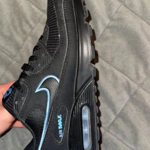 Svarta Nike Air Max 90 - Säljer ett par svarta Nike Air Max sneakers med blå detaljer och logga. Skorna har en synlig Air Max-sula och klassisk snörning. Perfekta för dig som gillar sportig stil och vill ha bekväma skor med snygg design. Snabb affär 