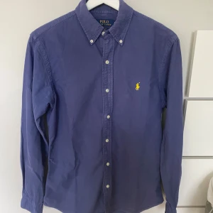 Polo Ralph Lauren skjorta  - Säljer en klassisk blå skjorta från Polo Ralph Lauren i slim fit-modell. Skjortan har långa ärmar, knappar framtill och den ikoniska gula broderade loggan på bröstet. Perfekt för dig som gillar stilrena plagg med snygga detaljer.hör av er vid intresse!!