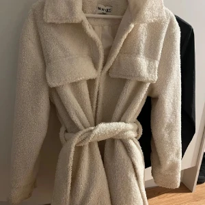 Beige teddyjacka med bälte från NA-KD - Mysig beige teddyjacka från NA-KD med krage, stora bröstfickor och knytskärp i midjan. Jackan har lång ärm och är perfekt för kyligare dagar. Stängs med bälte för en avslappnad look.