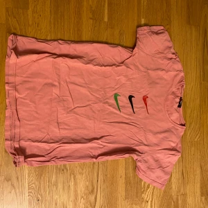 Rosa Nike t-shirt med färgglada swoosh - Säljer en rosa t-shirt från Nike med fyra swoosh-loggor i grönt, svart, rött och orange på bröstet. T-shirten har rund hals och korta ärmar. Perfekt för en sportig och avslappnad stil.