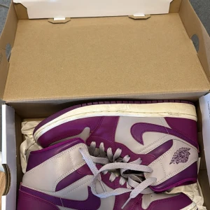 Nike Air Jordan 1 Mid Amethyst Ash/Red Plum - Säljer ett par Nike Air Jordan 1 Mid i färgen Amethyst Ash/Red Plum. Skorna har en snygg kombination av lila och ljusgrått med klassisk Air Jordan-logga på sidan. De har snörning och en vit sula som ger en sportig look. Perfekta för dig som vill sticka ut med dina sneakers.