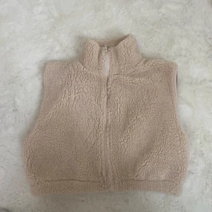 Beige teddyväst från NLY Trend - Mysig beige teddyväst från NLY Trend med hög krage och dragkedja framtill. Västen är croppad och har ett fluffigt, fleece-liknande material som ger en trendig look. Perfekt att slänga över en hoodie eller långärmad tröja.