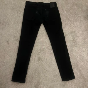 Svarta Replay Hyperflex jeans - Säljer ett par svarta Replay Hyperflex jeans med modellen anbass med klassisk femficksdesign och raka ben. Jeansen har en snygg svart läderpatch bak i midjan och är tillverkade i stretchigt material för extra komfort.