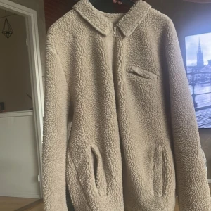 Beige teddyjacka med dragkedja - Mysig beige teddyjacka med dragkedja framtill och två fickor på sidorna. Jackan har en klassisk krage och är långärmad. Perfekt för kyliga dagar när du vill vara både varm och stilren.