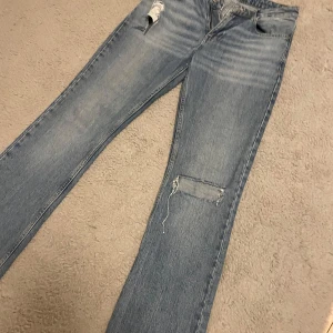 Blå slitna bootcut jeans - Säljer ett par blå bootcut jeans med slitningar på låret och knät. Jeansen är bootcut och lågmidjade. Perfekta för dig som gillar en avslappnad och trendig look.