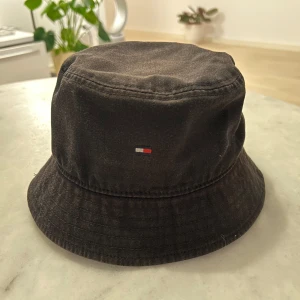 Svart bucket hat från Tommy Hilfiger - Snygg svart bucket hat från Tommy Hilfiger med diskret logga framtill. Hatten är i bomull och har klassisk form med nedvikt brätte. Perfekt accessoar för en stilren look. färgen bleknat