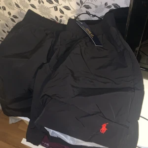 Svarta bad shorts från Polo Ralph Lauren - Snygga svarta shorts från Polo Ralph Lauren med röd logga broderad på benet. Elastisk midja med röd insida och meshfoder för extra komfort. Perfekta för sommardagar och har en klassisk, sportig look. Passar folk som har M också 