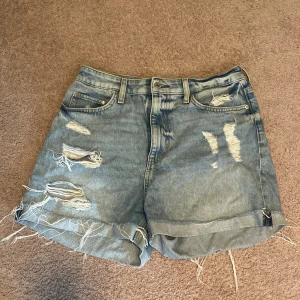 Blå slitna jeansshorts med fransar - Säljer ett par blå jeansshorts med slitna detaljer och fransig nederkant. Shortsen har klassisk femficksdesign, hög midja och uppvikta benslut. Perfekta för sommaren och ger en cool, avslappnad look.