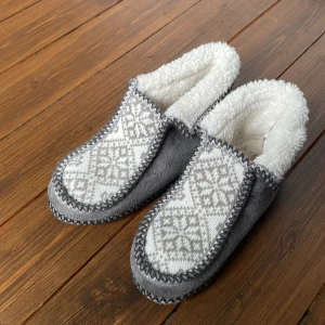 Grå och vita tofflor från Exani - Mysiga grå tofflor från Exani med vit, fluffig insida och stickat mönster på ovansidan. Perfekta för kalla dagar hemma. Snygg kontrastsöm runt kanterna och mjuk, vadderad insida för extra komfort.