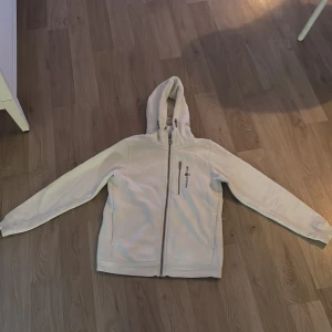 Vit fleecejacka med huva från Sail Racing - Säljer en vit fleecejacka från Sail Racing med huva och dragkedjor framtill. Jackan har en bröstficka med dragkedja och broderad logga på bröstet. Perfekt för kyliga dagar och enkel att matcha med andra plagg. 