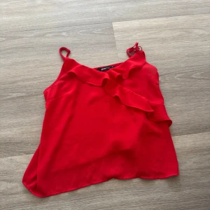 Röd volangtop med smala axelband - Säljer en snygg röd topp med smala axelband och volangdetalj framtill. Perfekt för att liva upp outfiten med en pop av färg. Lätt och luftig modell som passar till många tillfällen.