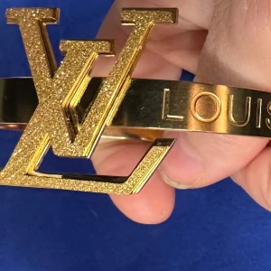 Guldigt supersnyggt armband . - Säljer ett stilrent armband från L  V i guldigt metallmaterial med stor glittrig -logga framtill. Armbandet har en bred design och tydlig text ingraverad längs bandet. Perfekt accessoar för att lyfta din outfit.