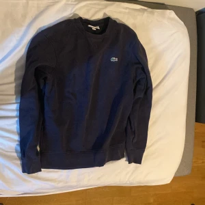 Mörkblå sweatshirt från Lacoste Sport - Säljer en mörkblå sweatshirt från Lacoste Sport med klassisk rund halsringning och den ikoniska gröna krokodilloggan broderad på bröstet. Tröjan har långa ärmar och är tillverkad i mjuk bomull. Perfekt för en sportig och stilren look. Passar för Grisch /laidback och  Orten stil.