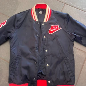 Bomberjacka från Nike - Snygg svart bomberjacka från Nike med rödvit ribbad krage, muddar och nederkant. Säljer för att den in tre har kommit till användning ❤️💙