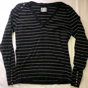 Svart v-ringad tröja med ränder från Esprit - Säljer en svart v-ringad tröja från Esprit med tunna vita ränder. Tröjan har långa ärmar och knappar både vid axeln och ärmsluten. 