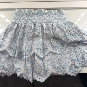 Vit och blå kjol med volanger - Charmig vit och blå kjol med ett detaljerat mönster och volanger. Den har en elastisk midja för bekväm passform. Perfekt för en somrig look! Endast använd en gång💘